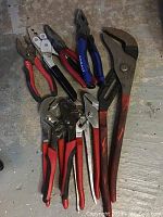 Photo showing a group of larger pliers and wrenches with red, black, and blue handles on a concrete floor.