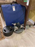 Blue foldable exercise mat leaning against furniture; assorted weight plates and dumbbell bars in front