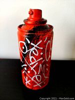 Front view of the spray can sculpture showing red base color with black and white painted graffiti-style markings.