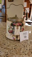 Front view showing floral design, silver plate lid with knob, and metal handle.