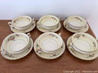 Six soup bowls arranged on matching side plates on a wooden surface showing floral pattern and gold trim