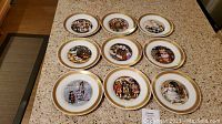 Nine decorative plates laid out on a table, each depicting a different illustrated scene with gold trim around the edges.