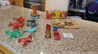 Wide view of vintage tin toys including red and green fighter planes, toy cars, tin tea set on tray, and boxed Airfix magnetic winder wheel, all on granite countertop.