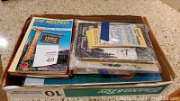 Box of assorted vintage atlases, maps and travel brochures showing colorful and black and white covers inside a large box