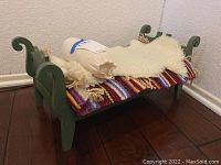 Toy bed in green wood with multicolored woven blanket, white faux sheepskin, and cream bolster pillow on top.