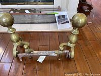 Pair of brass andirons on a wooden floor showing spherical finials and curved bases attached to iron bars.