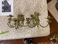 Top view of both brass French wall sconces showing three candles holders each and ornate scrolling arms placed on a beige floral tablecloth.