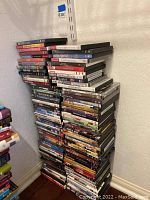 Large stack of assorted DVDs on floor against wall showing numerous titles and cases
