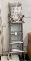 Folded aluminum Little Giant ladder with orange accents leaning against wall, hardware parts and manual attached in plastic bag