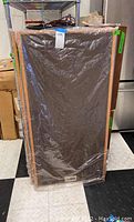 Front view of one speaker wrapped in plastic showing wood frame and black grill