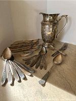 Overall view showing the set of spoons, forks, and knives with a large ornate silver-plated pitcher beside them on a stone counter.