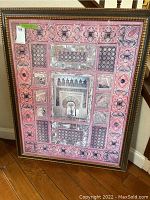 Framed wall art showing a pink border with geometric and floral motifs, central panel with architectural arch and palm trees