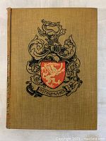 Front cover of the book showing tan cloth with a heraldic crest and Latin motto.
