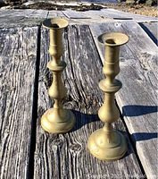 Pair of antique brass Victorian candlesticks photographed outdoors on a weathered wooden surface under natural daylight showing full view of items.