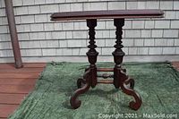 Full side view of antique table showing curved legs and turned pedestal supports, placed on green outdoor carpet with white shingled wall background.