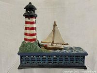 Front view of cast iron lighthouse with red and white stripes, green base and blue wave-patterned platform with sailboat beside it.