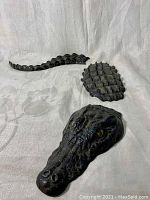 Three separate bronze pieces of alligator sculpture laid out on cloth background: head, mid-body, tail