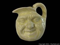 Front view of small ceramic pitcher sculpted as a man in the moon face on black background, showing detailed facial features with glossy pale yellow glaze.