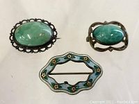Top view of three antique pins showing green stones and enamel decoration on different metal bases.