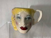 Front view of ceramic mug shaped as woman's face with yellow hair, red lips, and painted facial details.