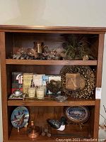 Wooden shelf unit with decorative artificial floral wreath, ceramic pitcher, books, decorative plates, candles, and figurines.