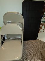 Five beige metal folding chairs stacked against a black folding table, showing the chairs' metal frame and smooth seats.