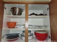 Wide shot of cupboard upper shelves showing colander, orange mixing bowl, CorningWare dishes, melamine bowls and lids, glass baking dishes, and soup bowls.