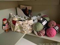 Wool yarn skeins in shades of green and pink, a knitted doll, and some crafting supplies on a shelf