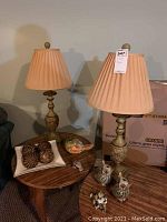 Two matching lamps with pleated beige fabric shades and decorative bases on a wooden table along with various decor items including owl figurines, wooden balls in a tray, bird and frog figurines, and a wooden platter.