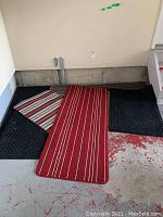 Seven assorted mats arranged on a concrete floor showing various colors and patterns including black textured mats, striped smaller mats, and one red runner mat.