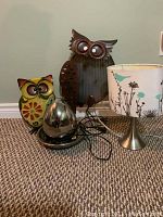 Photo of two metal decorative owls, a table lamp with painted bird and floral shade, and a small shiny silver fountain on a black base, all on carpeted floor.