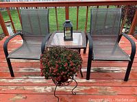 Full patio set showing two aluminum mesh chairs, glass top table, black lantern with candle on table, and metal plant stand with red natural flowers