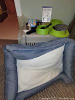 Photo showing dog bed in front with crate behind, on top of crate are green dog bowls and some pet accessories like grooming items and clicker training devices.