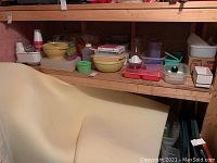 Shelf with multiple plastic food containers, bowls in yellow, green, and cream colors, plastic cups, Tupperware containers, and boxes.