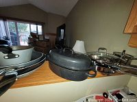 Shelf with various pots and pans including a black enamel roaster, several stainless steel pots with lids, and non-stick fry pans stacked together.