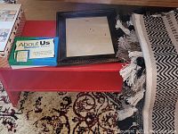 Lot on red shoe rack, showing About Us game, empty black photo frame, woven mat folded, and shoe rack surface