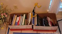 Two cardboard boxes filled with various books and a yellow twin hook style steering lock with key inserted in lock, placed on floor near wall with outlet.