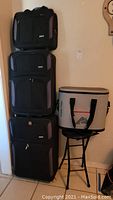 Stack of three black luggage suitcases with gray and purple accent panels, three sizes, standing upright side by side, cooler on top of collapsible stool