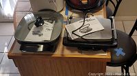 Hamilton Beach panini press on the right with Farberware electric skillet on the left, both on a wooden surface, item cords visible.
