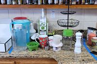 Photo of kitchen counter showing two plastic pitchers, glass pitcher, various cups, white slicer, manual green chopper, white pepper grinder, and black three-tier metal rack