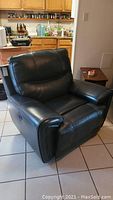 Front angled view of black leather electric recliner showing padded armrests, cushioned backrest, and overall shape.