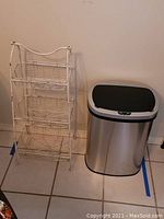 Photo of white metal 3-tier stand with three wire baskets, next to the trash can to show size and design.