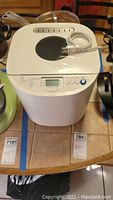Top front view of Oster bread maker showing digital control panel, lid with viewing window, and power cord attached.