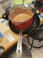 Red copper pot with glass lid and strainer on top of stove, showing handle and locking mechanism.