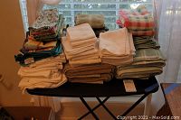 Full view of multiple stacks of towels on a table by window, showing various colors and sizes including terry cloth and printed towels.