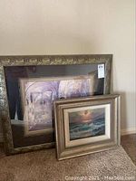 Photo showing both framed art pieces leaning against a wall, one larger print with dark ornate frame and one smaller seascape painting with lighter frame.