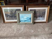 All three framed artworks arranged on carpeted stairs showing two city street scenes and one ocean wave scene.