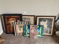 View of all seven artworks grouped on floor against wall showing mix of framed and unframed pieces with varied artistic themes including floral, abstract, architectural, and surrealistic art.