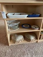 Photo of white and blue ceramic baking dishes on top shelf, floral patterned plates and wire shelf organizer on middle shelf, heart-shaped cushions on bottom shelf of wooden shelving unit.