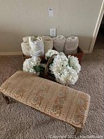 View of the upholstered bench stool, basket of faux flowers in front, and basket of rolled towels behind on carpet.